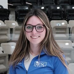 Portrait of a smiling woman with long light brown hair and pink glasses, wearing a blue polo shirt, standing in an indoor arena with seating behind her.