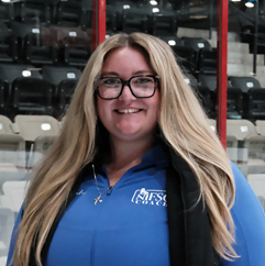 Smiling woman with long blonde hair and glasses, wearing a blue polo with a logo, in an arena.
